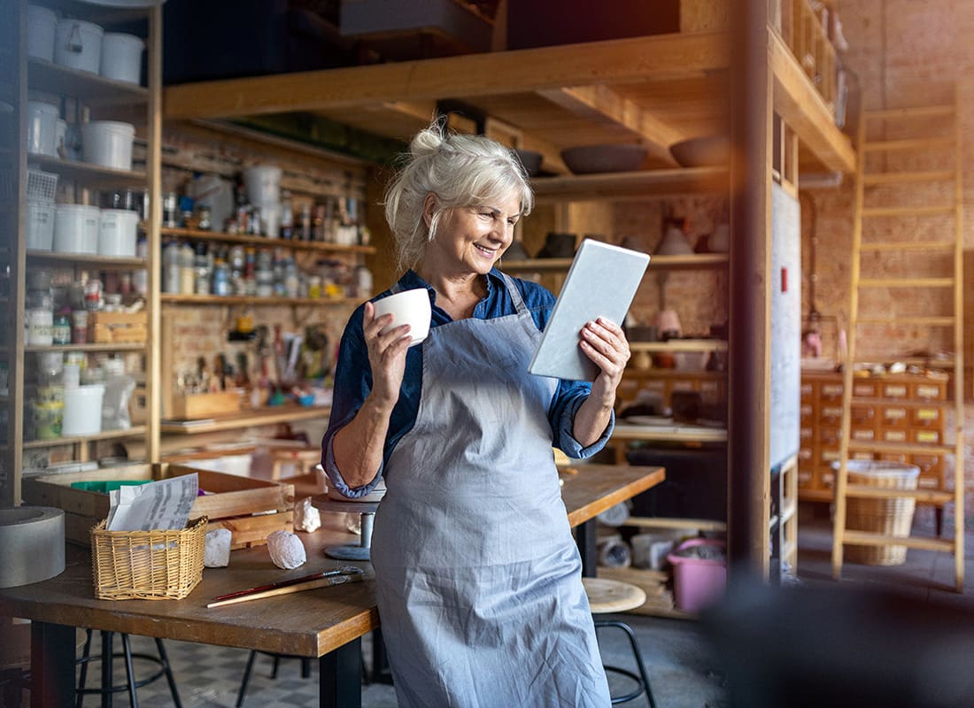 Business Insurance - Portrait of a Cheerful Elderly Woman Holding a Tablet and Coffee Mug Standing in her Arts and Crafts Shop
