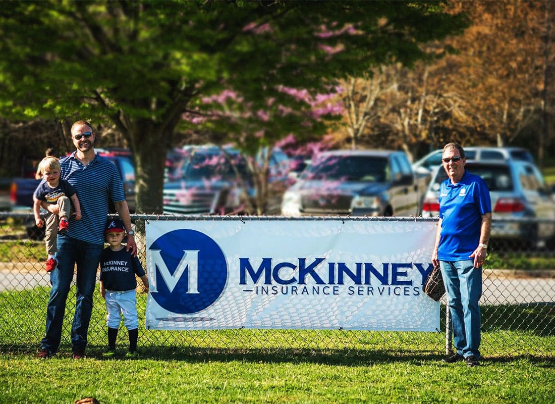 Refer Us - Portrait of Chad McKinney with his Two Sons Standing Next to a McKinney Insurance Sign on a Baseball Field on a Sunny Day